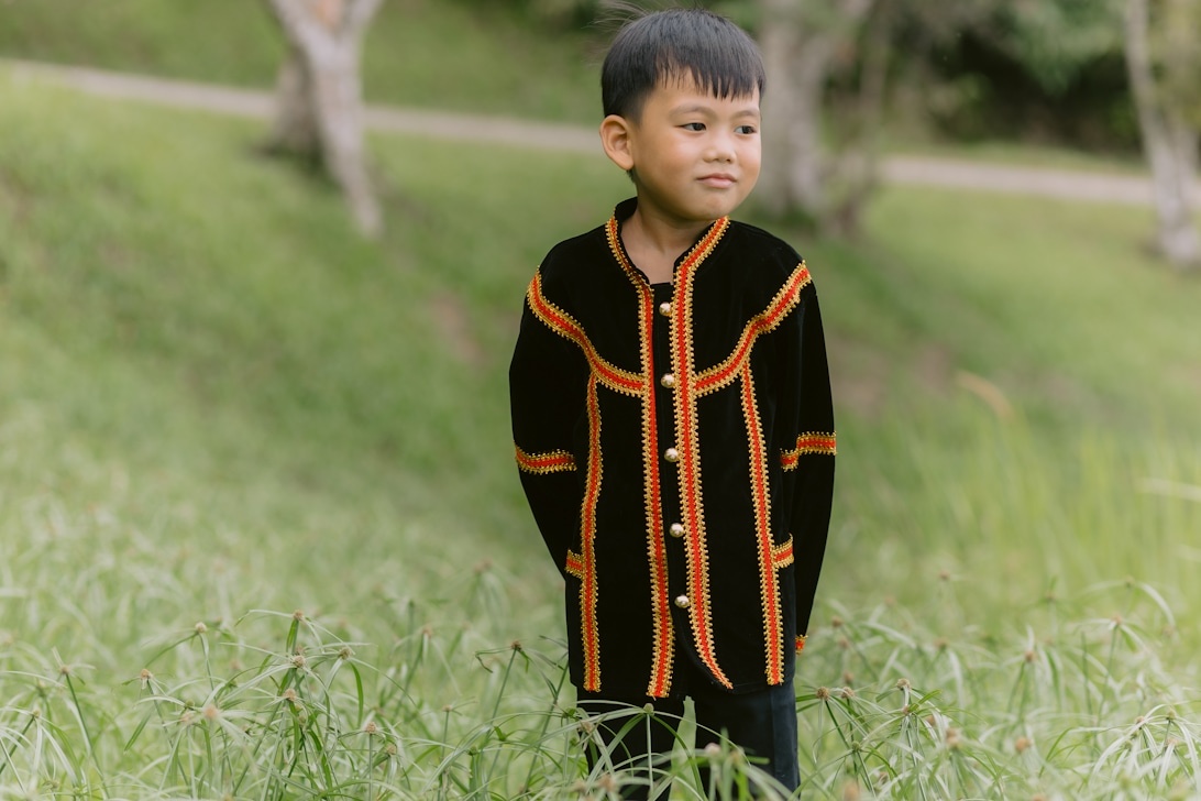 Ein kleiner Junge in einer eleganten, zeremoniell anmutenden schwarzen Jacke mit orangefarbenen und roten Verzierungen, die sich perfekt für einen stilvollen Burgbesuch eignet.